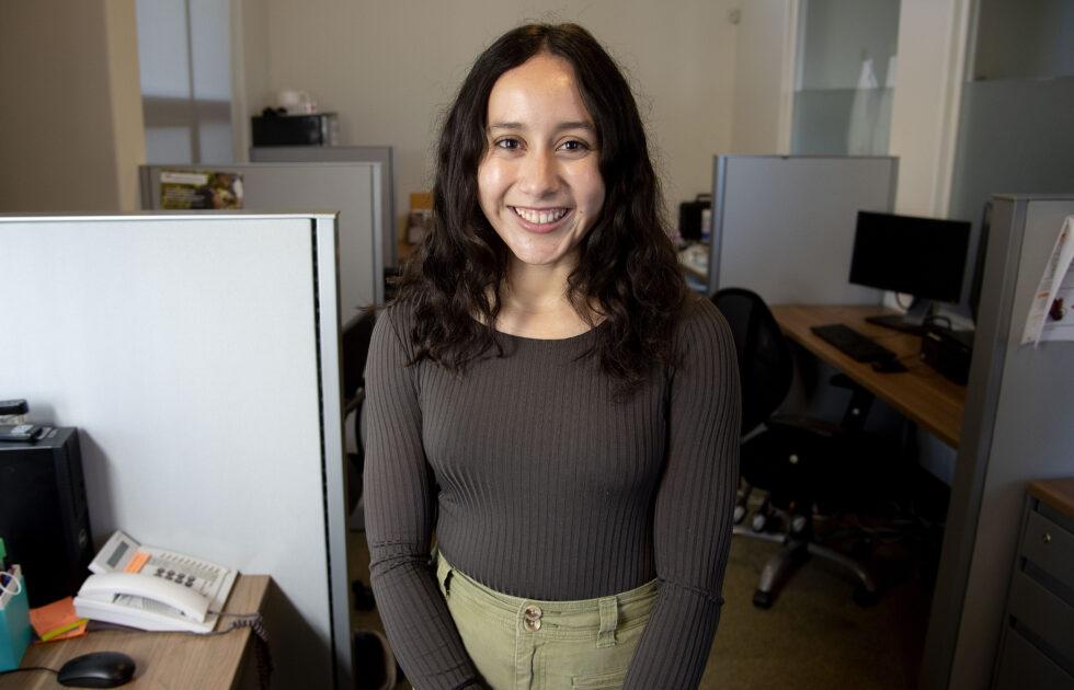 Young professional woman standing and smiling in an office cubicle workspace environment.