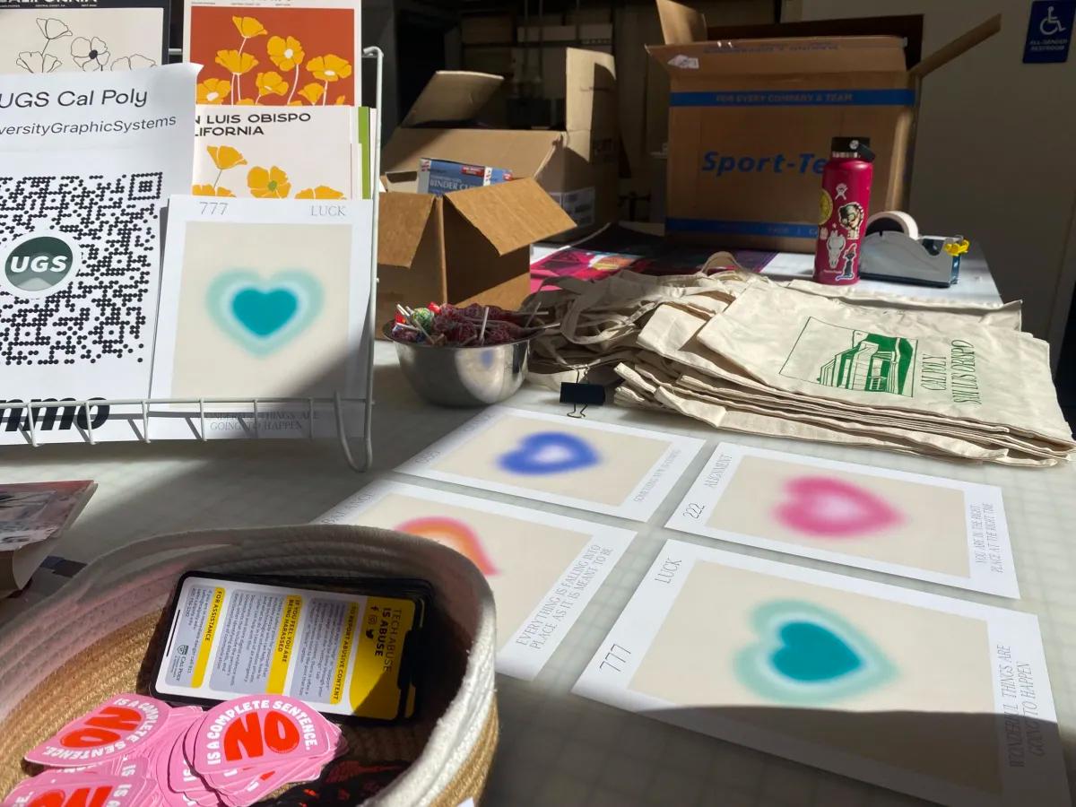 A display table for University Graphic Systems at Cal Poly featuring colorful heart prints, tote bags, stickers, and postcards promoting student-designed artwork and printing services.