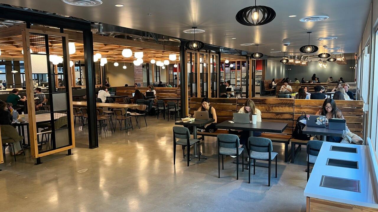 Students study and eat inside Cal Poly’s 1901 Marketplace dining area, featuring modern wood and metal decor, hanging lights, and open seating with tables and booths.