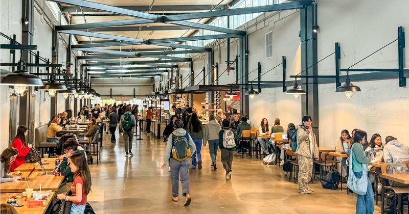 Students and visitors sit and walk through a bright, industrial-style dining hall with exposed beams, pendant lighting, and long wooden tables inside Cal Poly’s 1901 Marketplace.