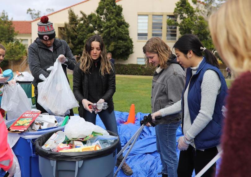 Students participate in a campus waste reduction event, sorting trash and recyclables into bins on a grassy area.