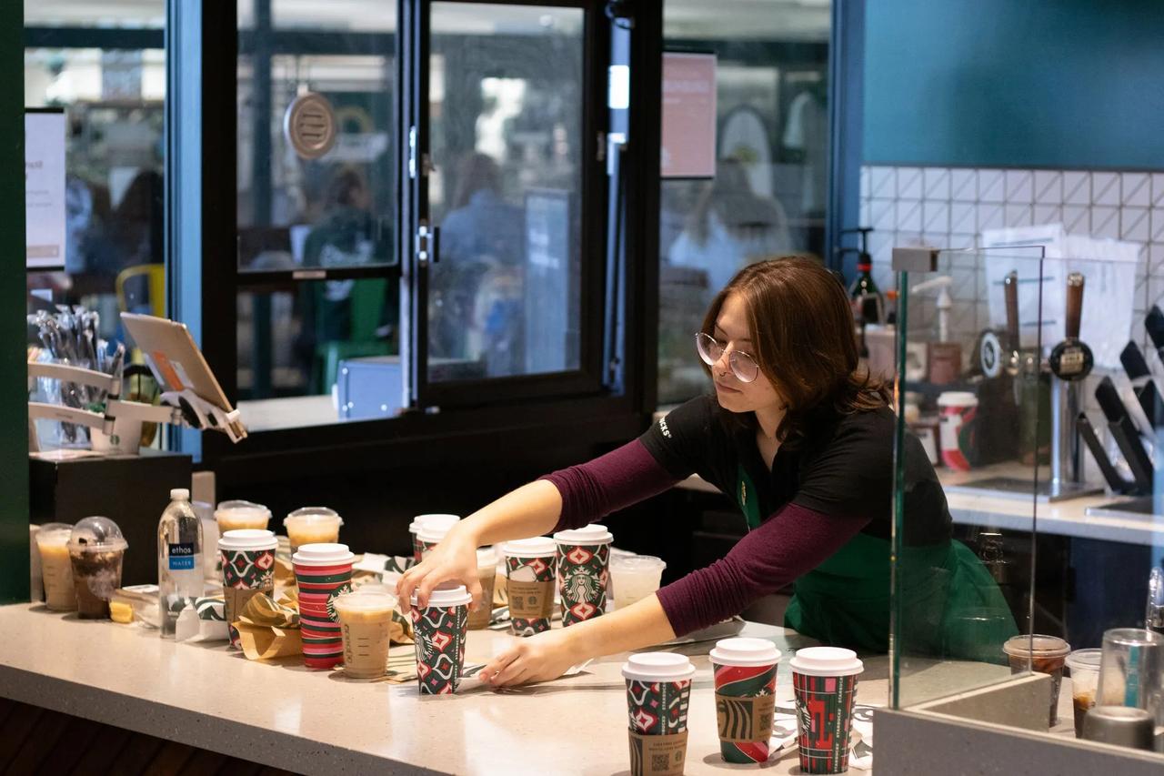 Barista arranges multiple Starbucks drinks on the counter, surrounded by festive holiday cups and café equipment.