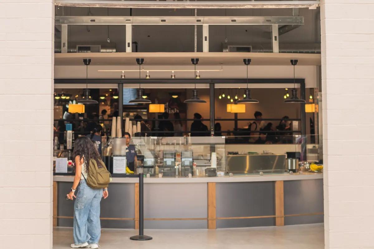 Student with a backpack orders at a modern campus café counter with hanging lights and baristas working behind glass.