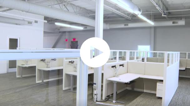 Bright modern office space with white cubicles, glass partitions, and exposed ceiling ducts under fluorescent lighting.