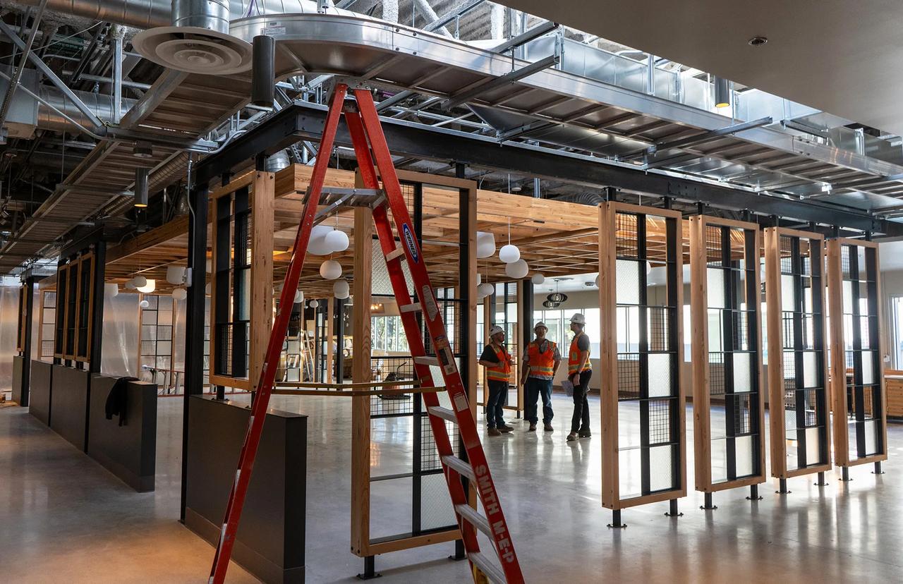 Construction workers in safety vests and helmets stand inside a modern building under construction with exposed ductwork, wooden beams, and a red ladder in the foreground.