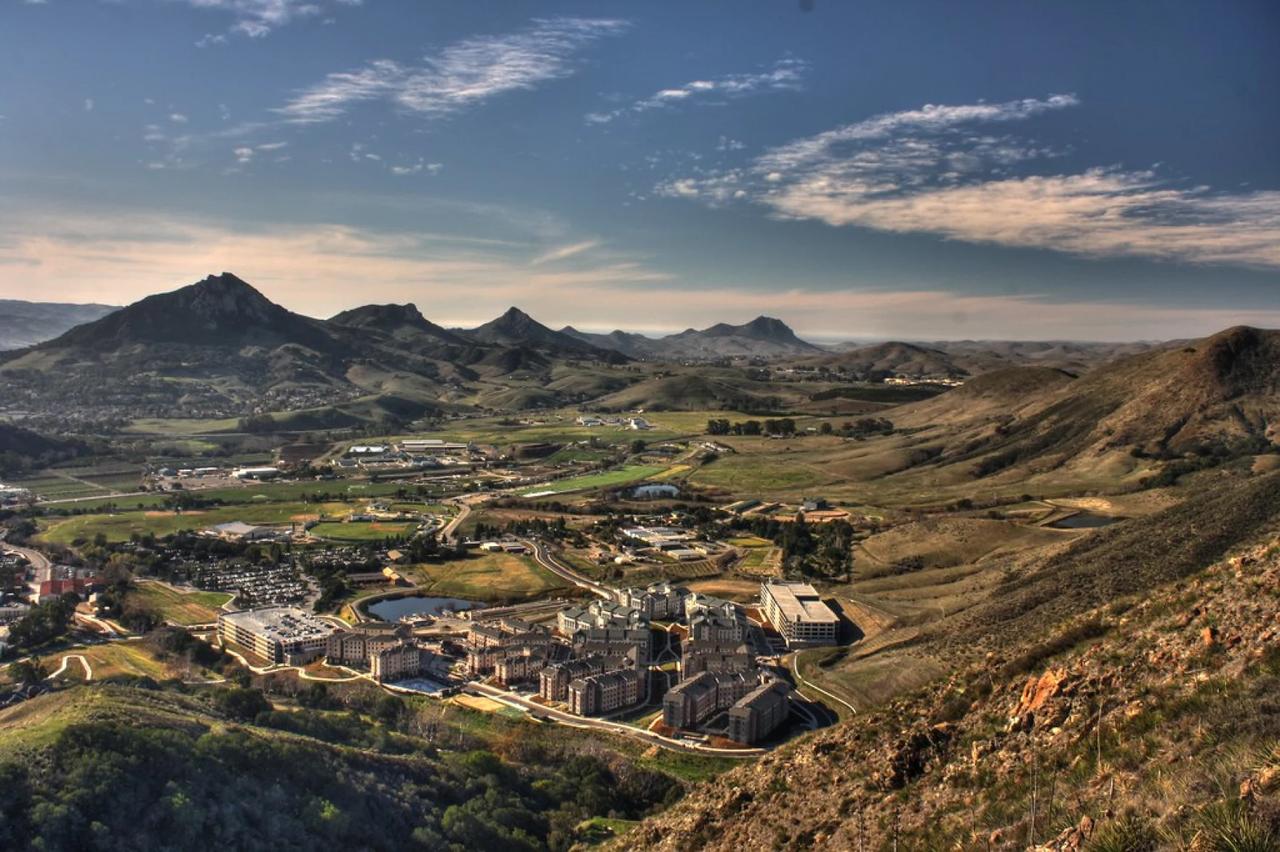 Aerial view of Cal Poly San Luis Obispo nestled in a valley surrounded by rolling green hills and the Nine Sisters volcanic peaks under a partly cloudy sky.
