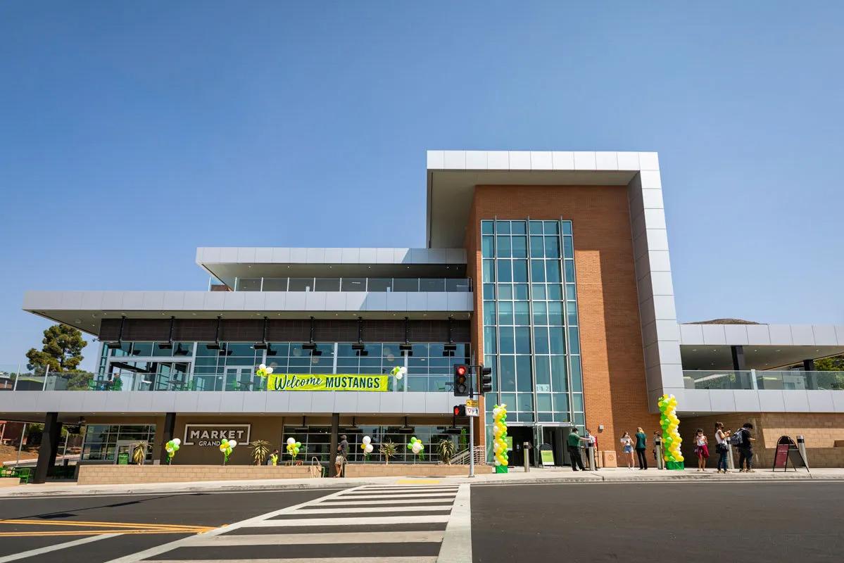 Exterior view of the Vista Grande dining complex at Cal Poly, featuring modern glass and brick architecture, green and gold balloons, and a banner reading “Welcome Mustangs.”