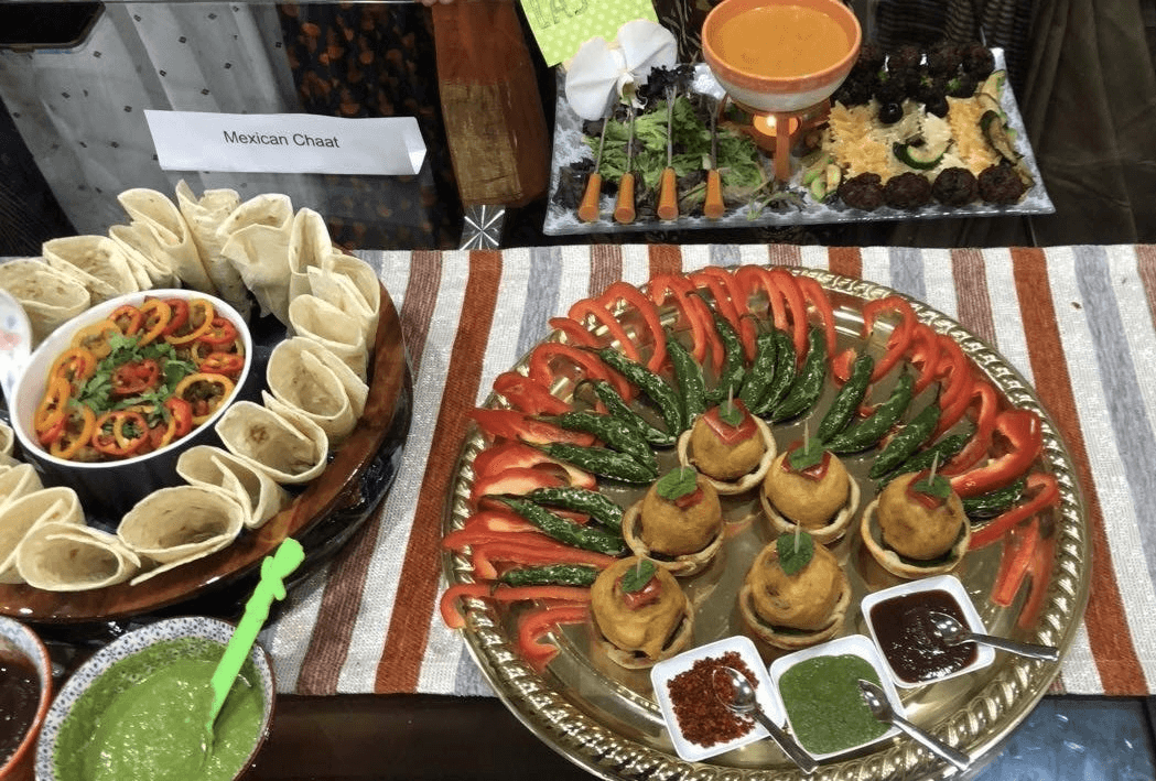 Colorful food display labeled “Mexican Chaat,” featuring rolled tortillas, sliced peppers, fried appetizers with mint garnish, green and red chutneys, and a striped tablecloth beneath the platters.