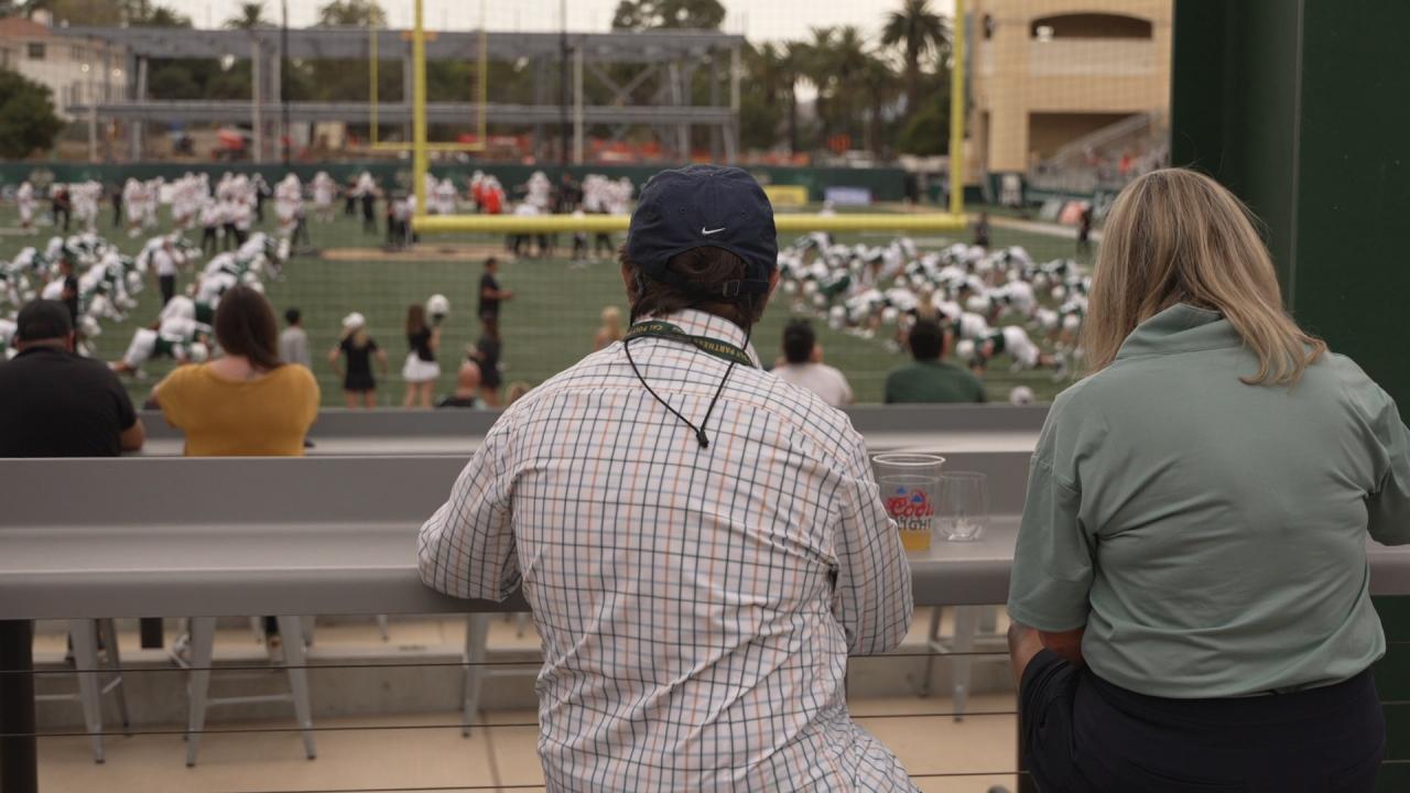 Two people sit at an outdoor counter overlooking Cal Poly’s football field as players warm up on the turf, with fans and cheerleaders visible in the background.