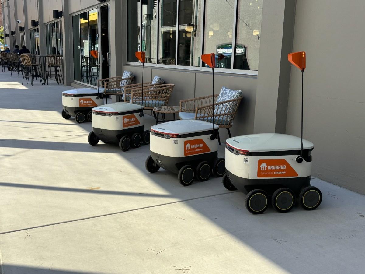 Row of four Grubhub delivery robots parked outside a building with wicker chairs and tables, ready for use on a sunny day.