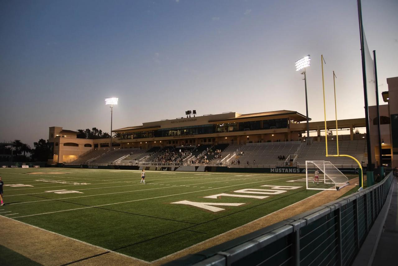 Evening view of Cal Poly’s Alex G. Spanos Stadium illuminated by field lights, with a few players on the turf and spectators seated in the stands under a twilight sky.