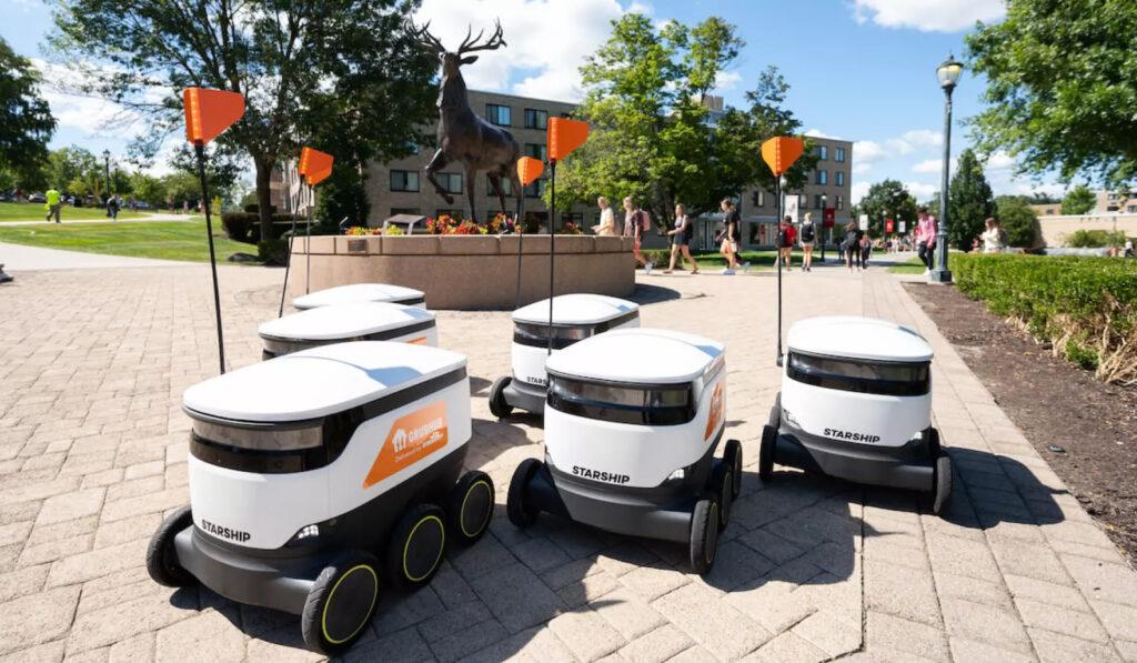 Group of Grubhub Starship delivery robots with orange flags gathered on a university walkway near a bronze deer statue, with students walking in the background on a sunny day.
