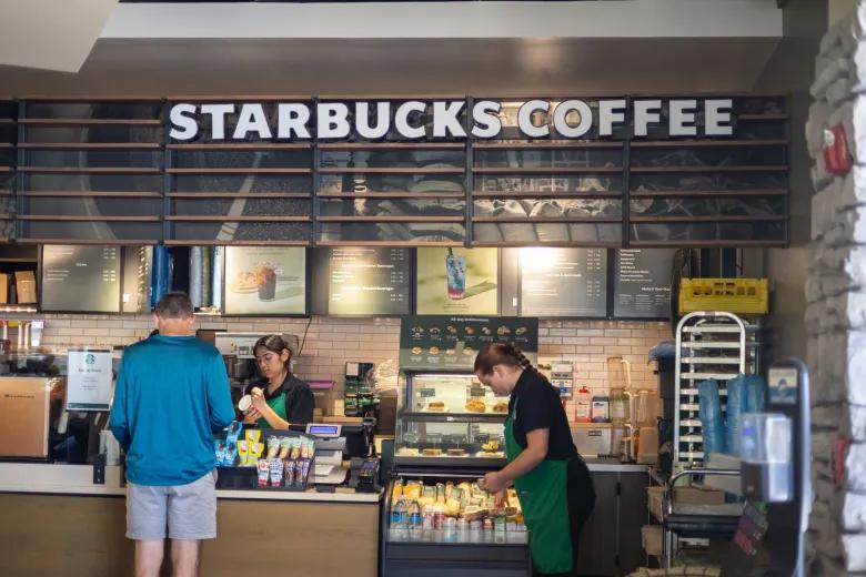Two Starbucks employees stand behind the counter serving a customer inside a Starbucks café, with the illuminated menu and “Starbucks Coffee” sign visible above.
