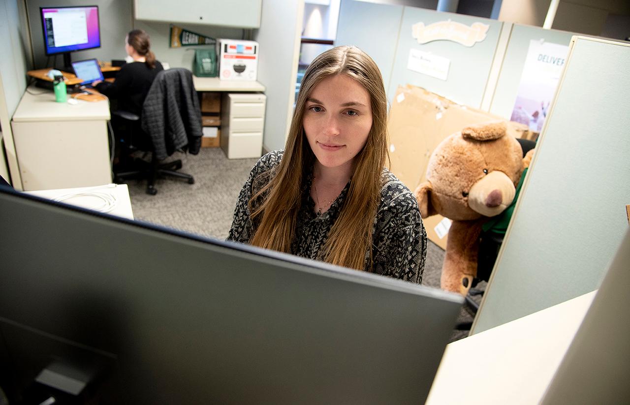 Woman working at her computer in an office cubicle with another coworker in the background and a large stuffed bear seated beside her desk.