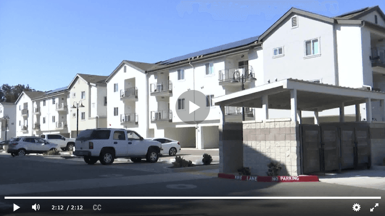 Still frame from a news video showing the exterior of the Harvest Lofts apartment complex in San Luis Obispo, featuring white multi-story buildings with balconies, parked cars, and a clear blue sky.