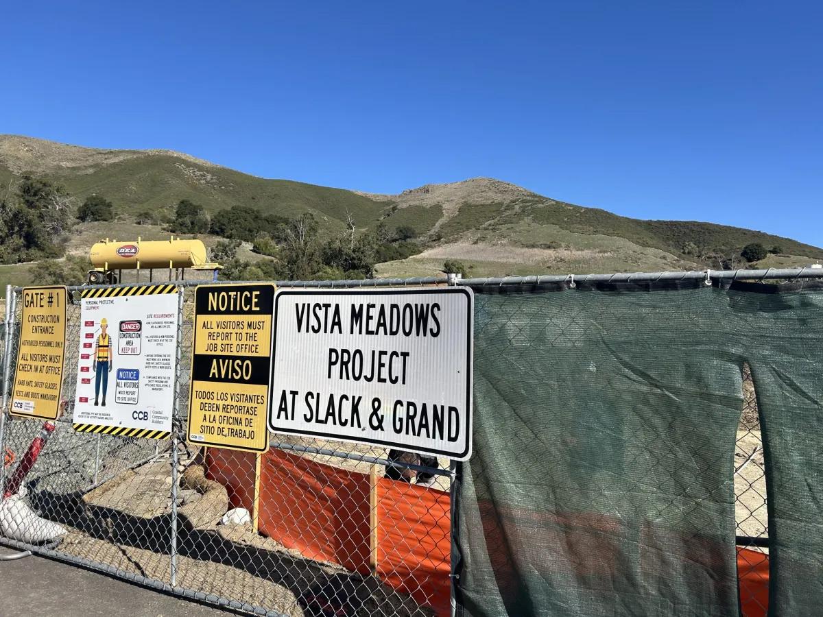 Construction site entrance at the Vista Meadows Project located at Slack and Grand in San Luis Obispo. Signs on a chain-link fence display safety notices in English and Spanish, with hills and a clear blue sky in the background.