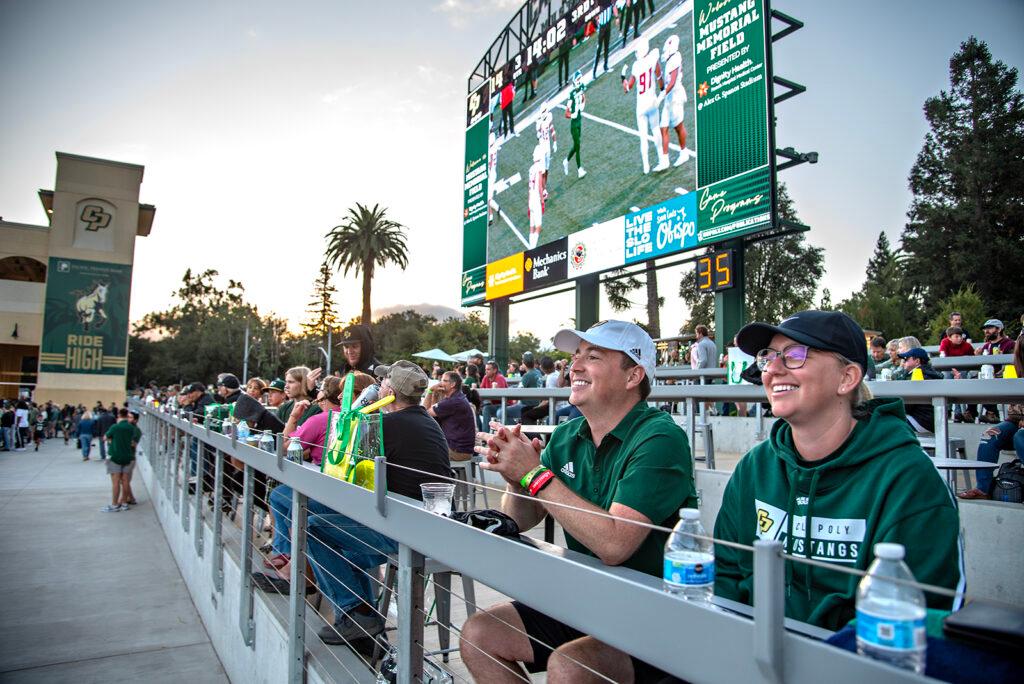 Fans sitting and smiling in the stands at Cal Poly’s Alex G. Spanos Stadium during a football game, with the scoreboard and large video screen visible in the background.