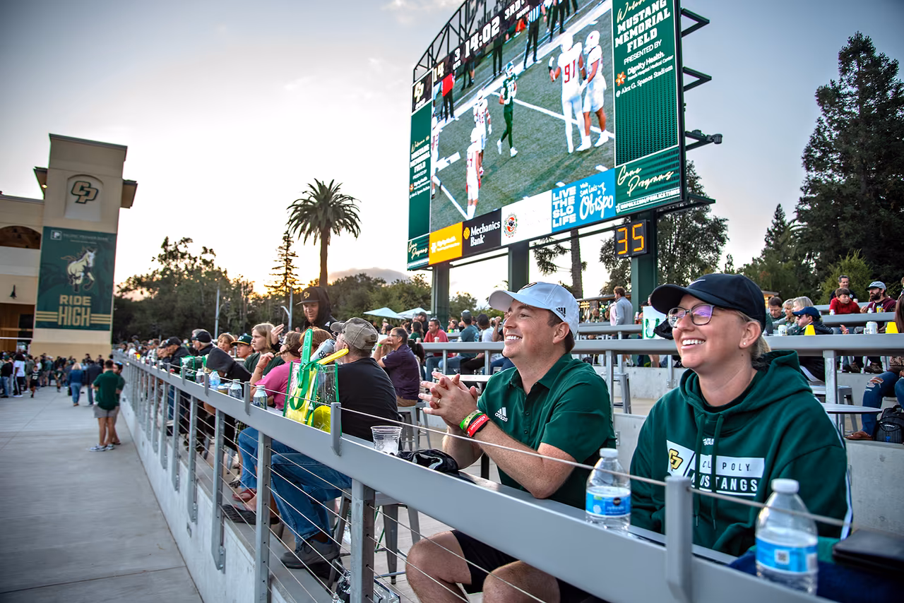 Fans sitting and smiling in the stands at Cal Poly’s Alex G. Spanos Stadium during a football game, with the scoreboard and large video screen visible in the background.