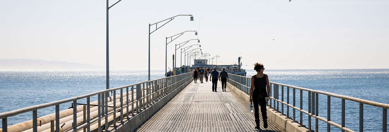 People walk along a long industrial pier stretching into the ocean under clear skies, with distant hills visible on the horizon and sunlight reflecting off the water.