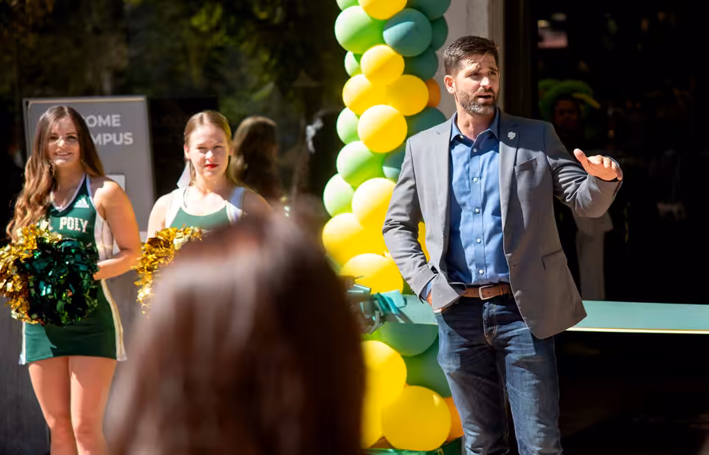 A man in a blazer speaks at a ribbon-cutting ceremony, flanked by Cal Poly cheerleaders and a green and yellow balloon arch in the background.