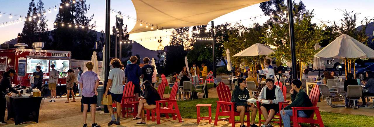 Students gather and dine outdoors in the evening near a food truck under string lights, with red Adirondack chairs and umbrella-covered tables creating a festive, relaxed atmosphere.