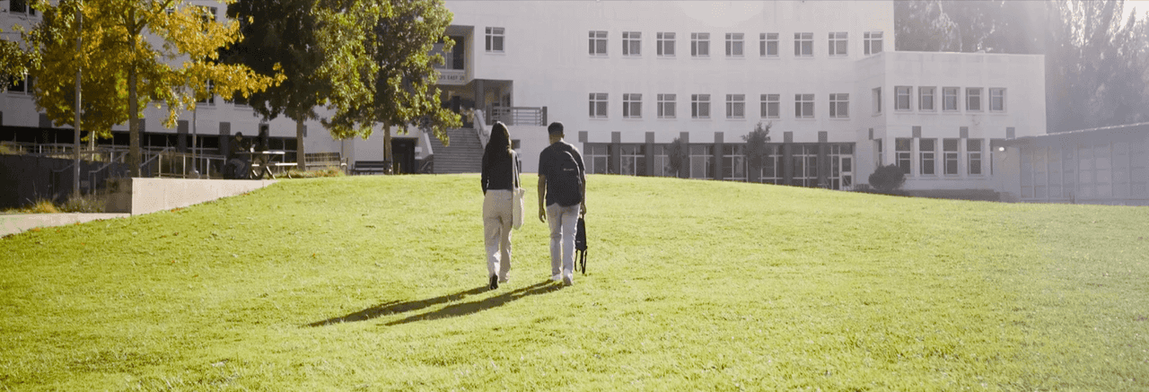 Two students walk side-by-side across a sunlit grassy lawn toward a modern academic building with large windows and trees showing fall colors.