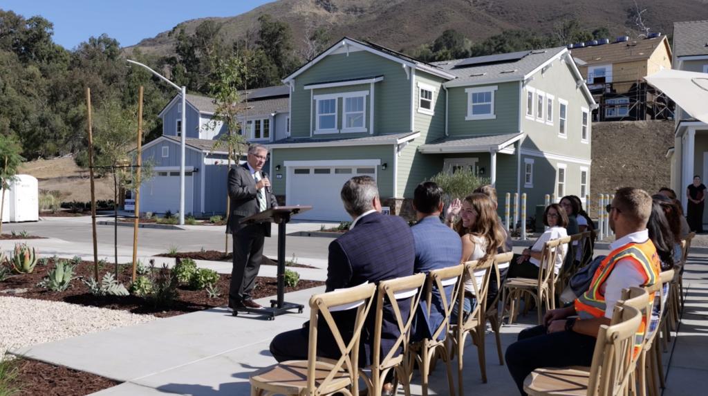 A speaker addresses attendees at the dedication ceremony for Cal Poly’s new faculty and staff housing neighborhood in San Luis Obispo, with newly constructed homes in the background.