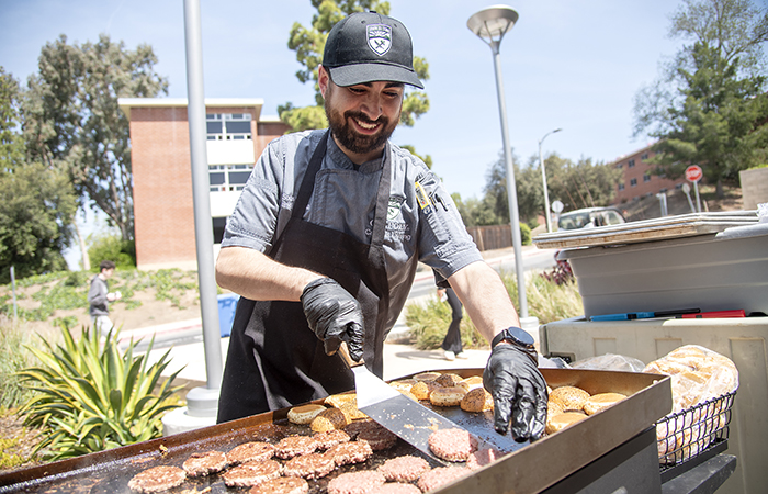 A Cal Poly Campus Dining staff member cooking hamburgers on an outdoor flattop grill.